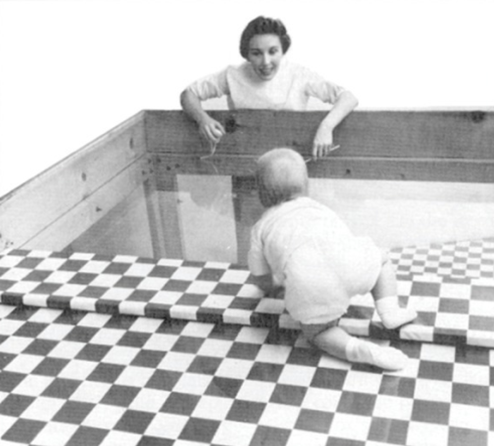 Een foto van een baby die richting een vrouw kruipt. De baby kruipt binnen een houten vierkante frameconstructie met glazen bodem waarvan de helft door een geblokt patroon bedekt is. De vrouw staat buiten het houten frame en nodigt de baby lachend uit om naar haar toe te kruipen. De baby is bijna halverwege en begint de doorzichtige helft van de ondergrond te bereiken.